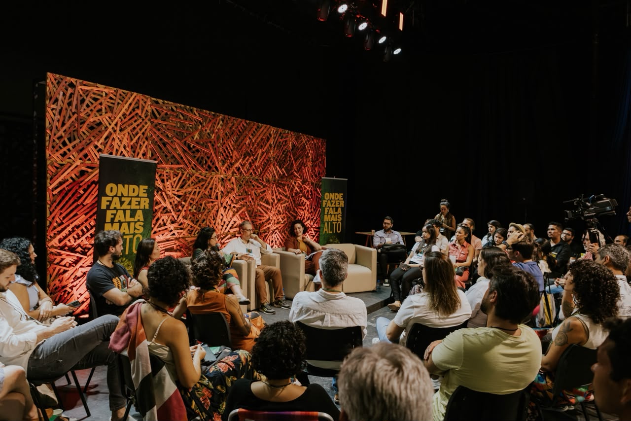 Mais de 80 mulheres que impulsionam a sociobiodiversidade amazônica ganham destaque no FIINSA 2026 Roda de conversa no FIINSA com três participantes em poltronas diante de painel de madeira laranja e público sentado ao redor